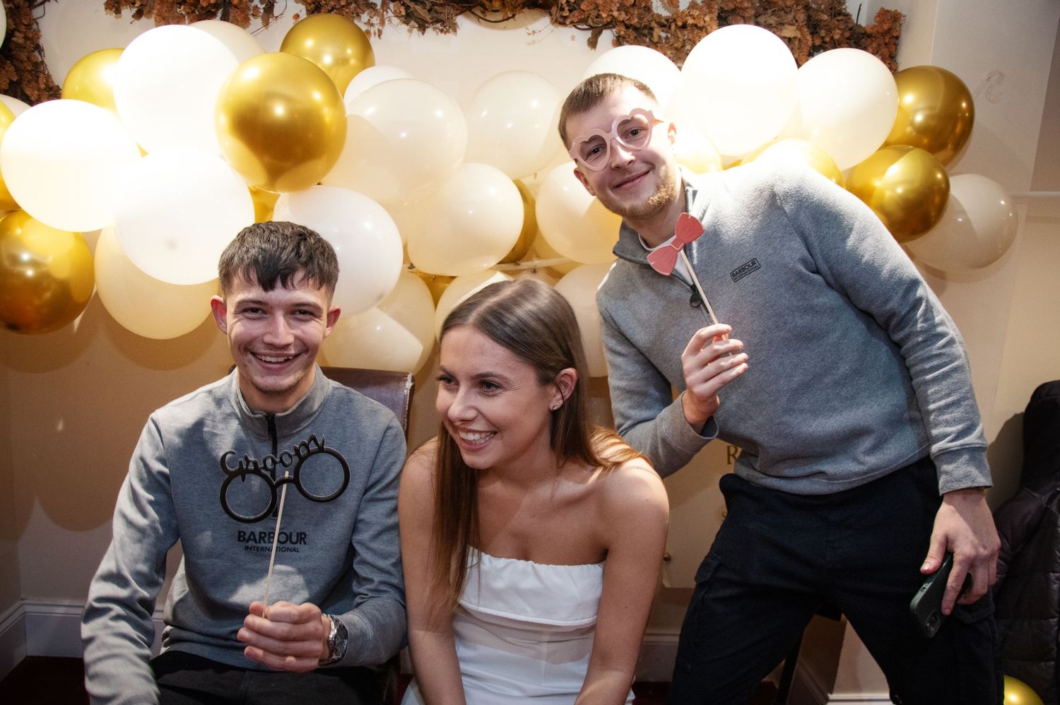 Party guests pose against a festive balloon wall backdrop with white and gold decorations.
