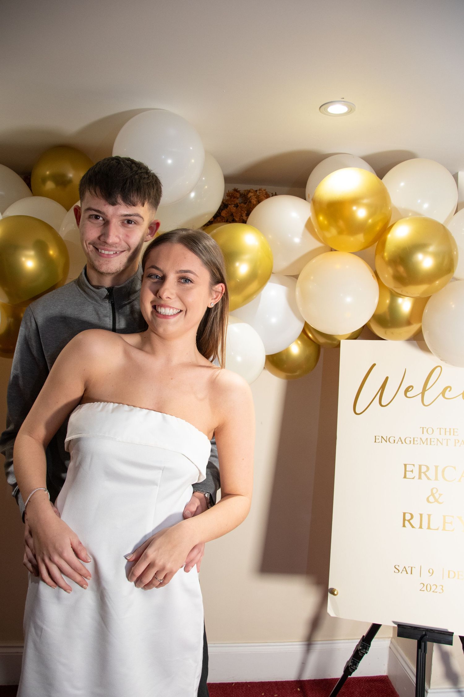 Couple poses in front of gold and white balloon arch at an engagement celebration event.