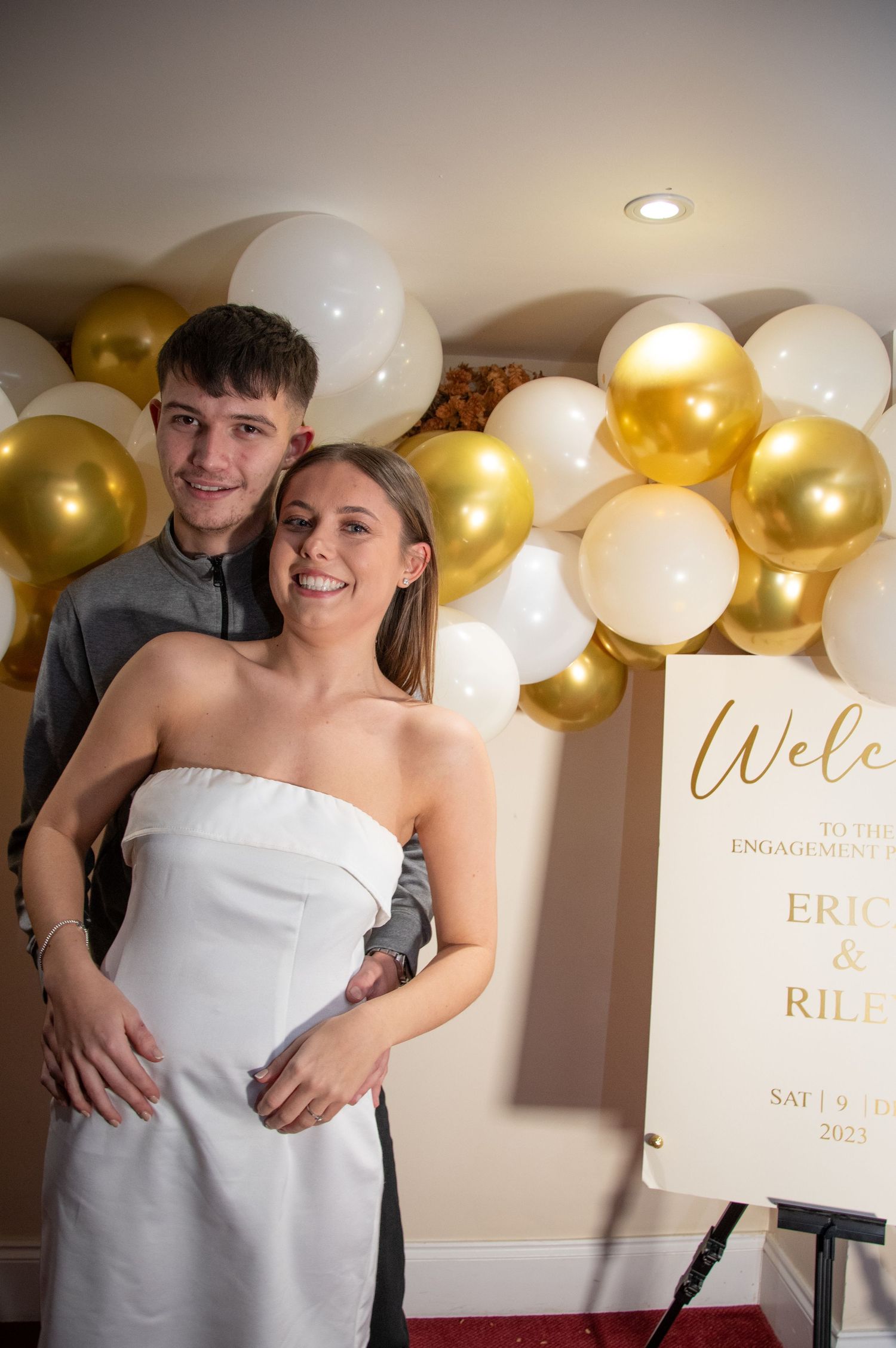 A couple embraces in front of a decorative gold and white balloon wall at their engagement celebration.