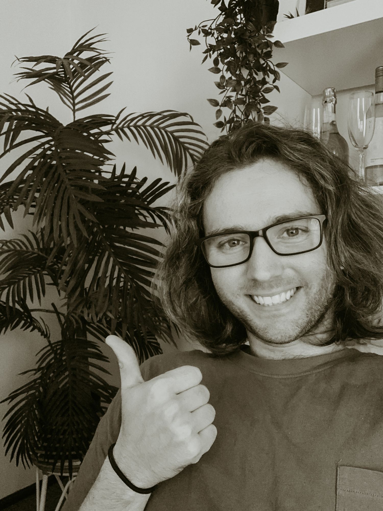 Black and white portrait of person with glasses giving thumbs up next to indoor plant.
