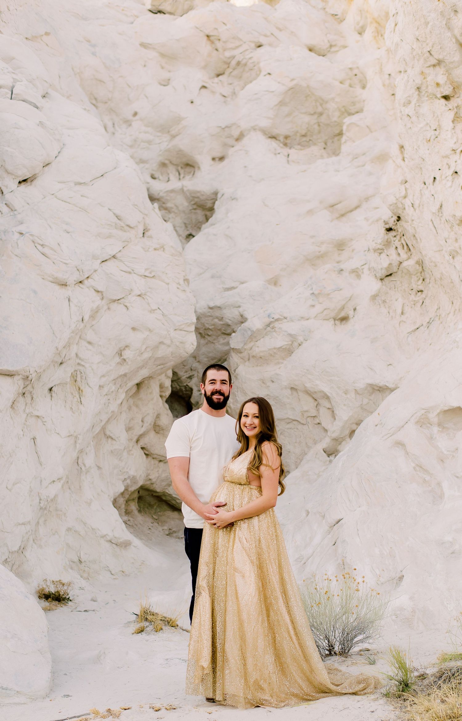 Couple posing in front of white rock formations, woman in gold gown, man in white shirt, maternity photo shoot.