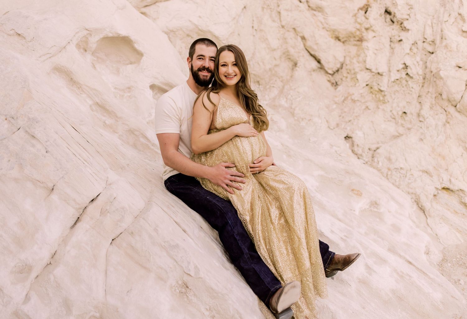 Couple poses on white rock formation, woman in gold maternity gown cradling baby bump, man in navy pants embracing her.