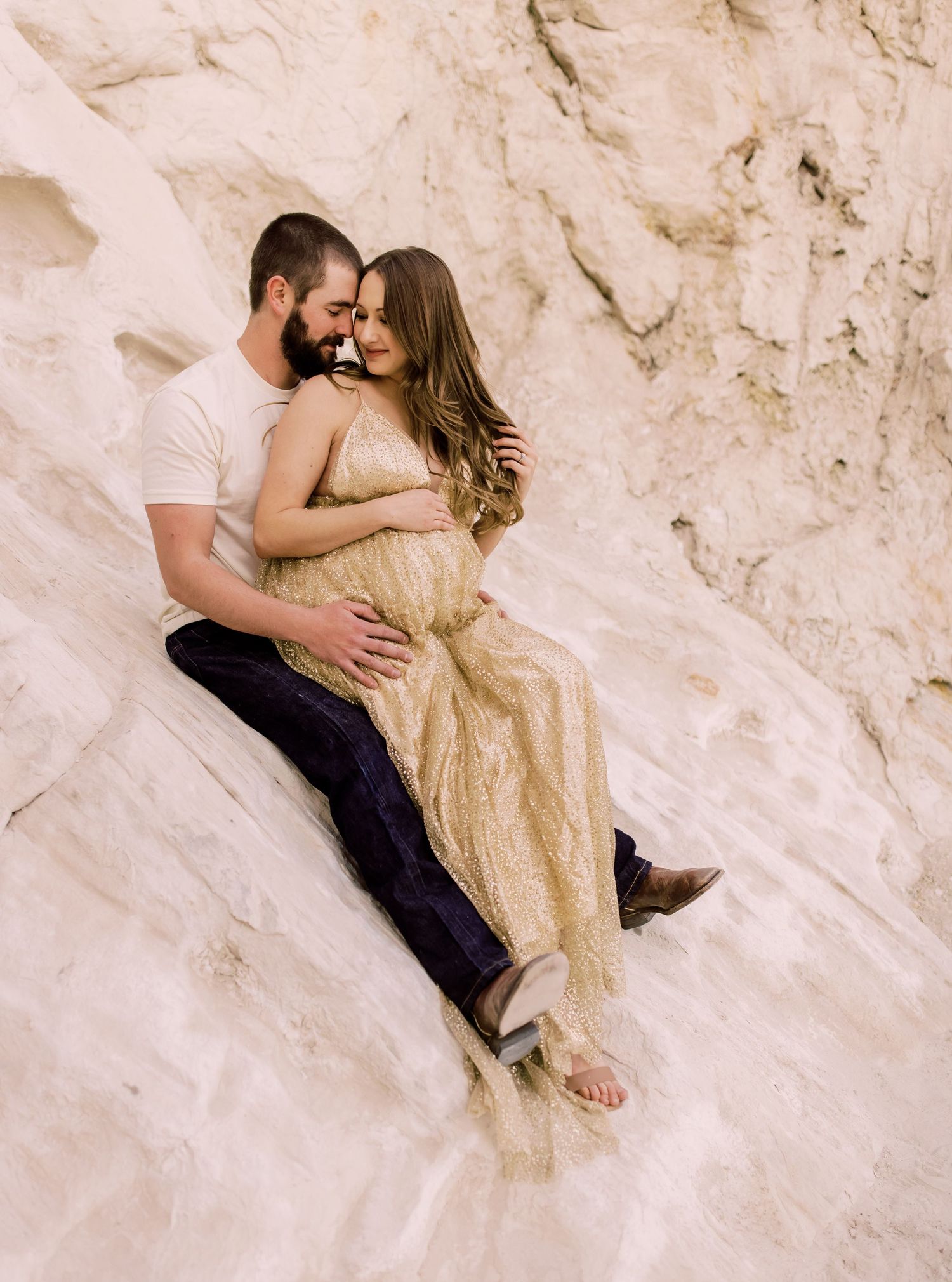 A couple sits intimately together on white rocks wearing casual attire and a flowing gold dress for a romantic photoshoot.