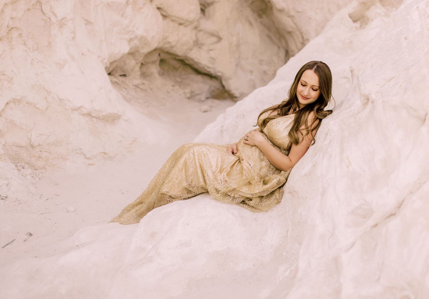 Pregnant woman in gold lace gown reclining on white sand dunes for a serene maternity photo shoot.