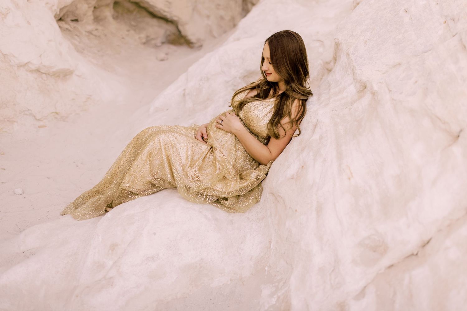 Pregnant woman in gold lace gown reclines gracefully on white sandy rocks during an outdoor maternity photo session.