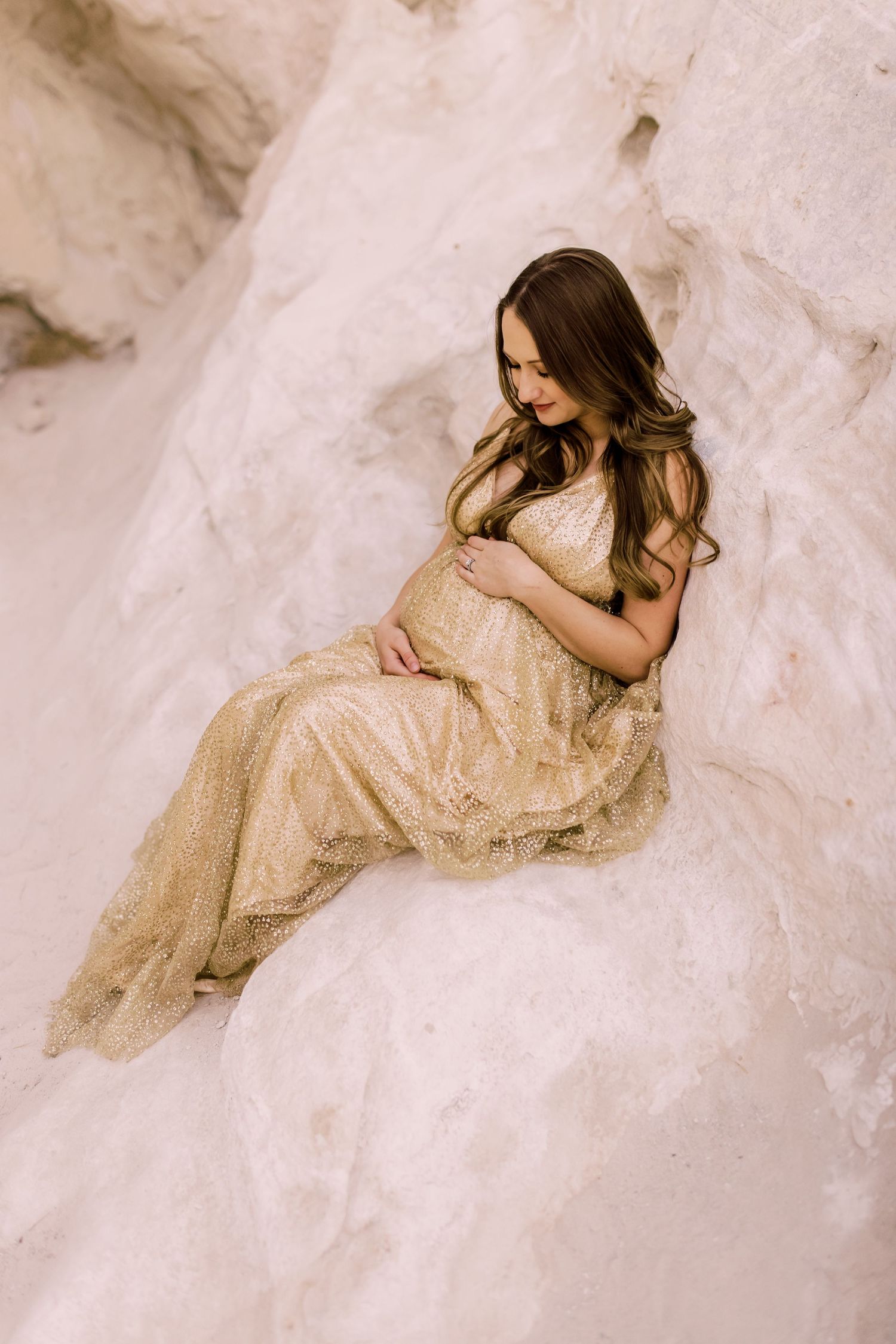 Expecting mother in flowing beige lace gown poses on white textured background during maternity photoshoot.