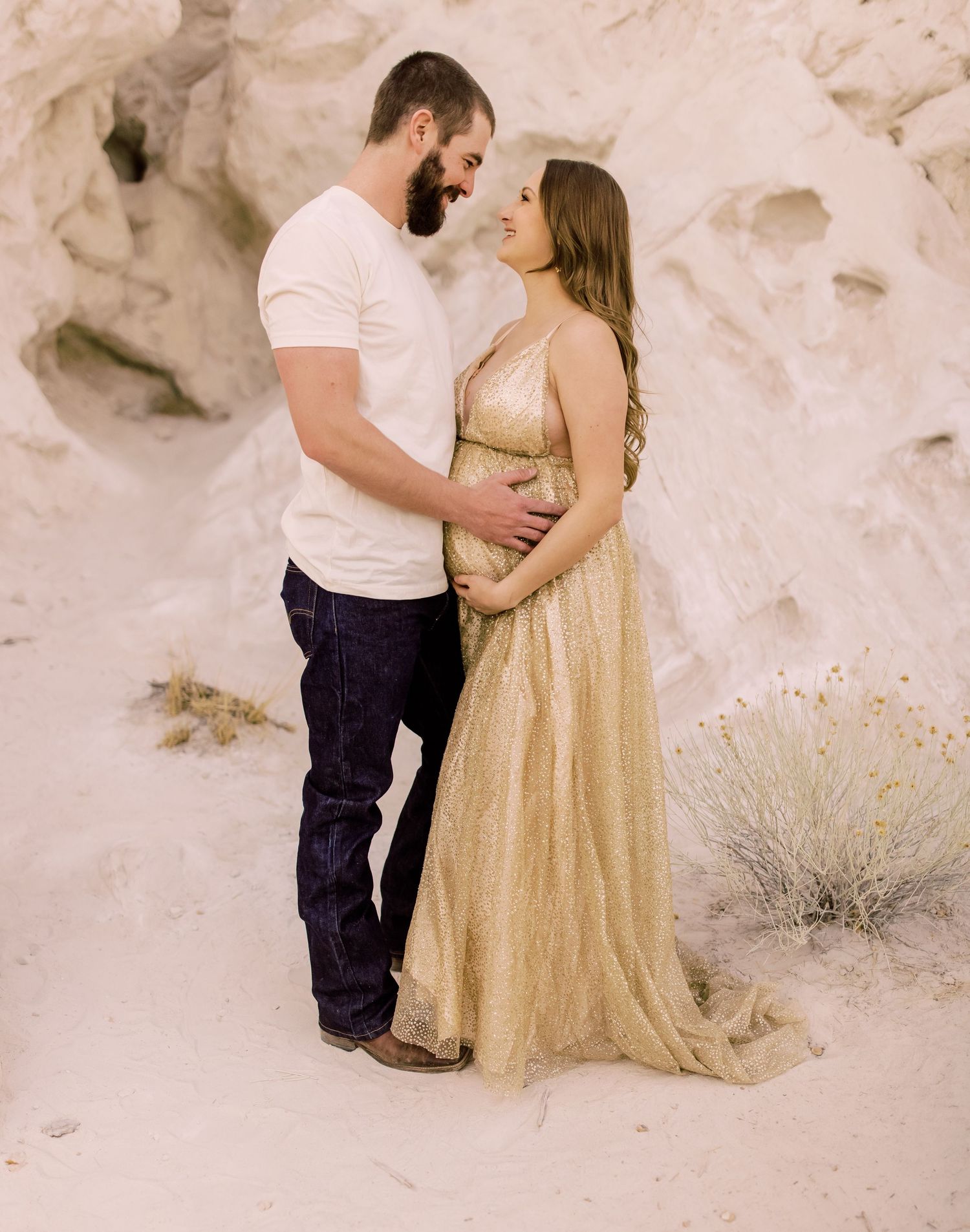 Couple in romantic desert engagement photo with woman wearing gold dress standing against white rock formation backdrop.