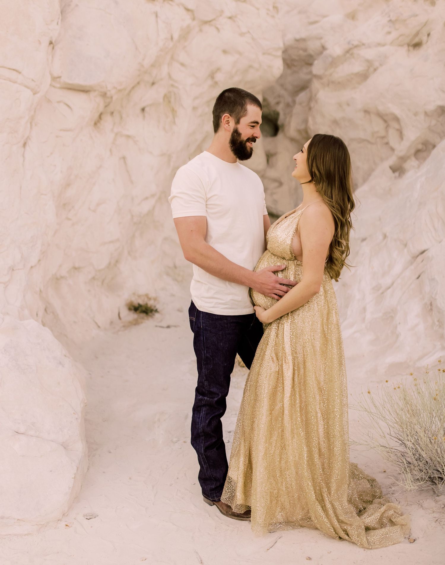 Pregnant couple poses in white rock canyon, woman in gold gown, man in white tee and jeans, cradling baby bump.