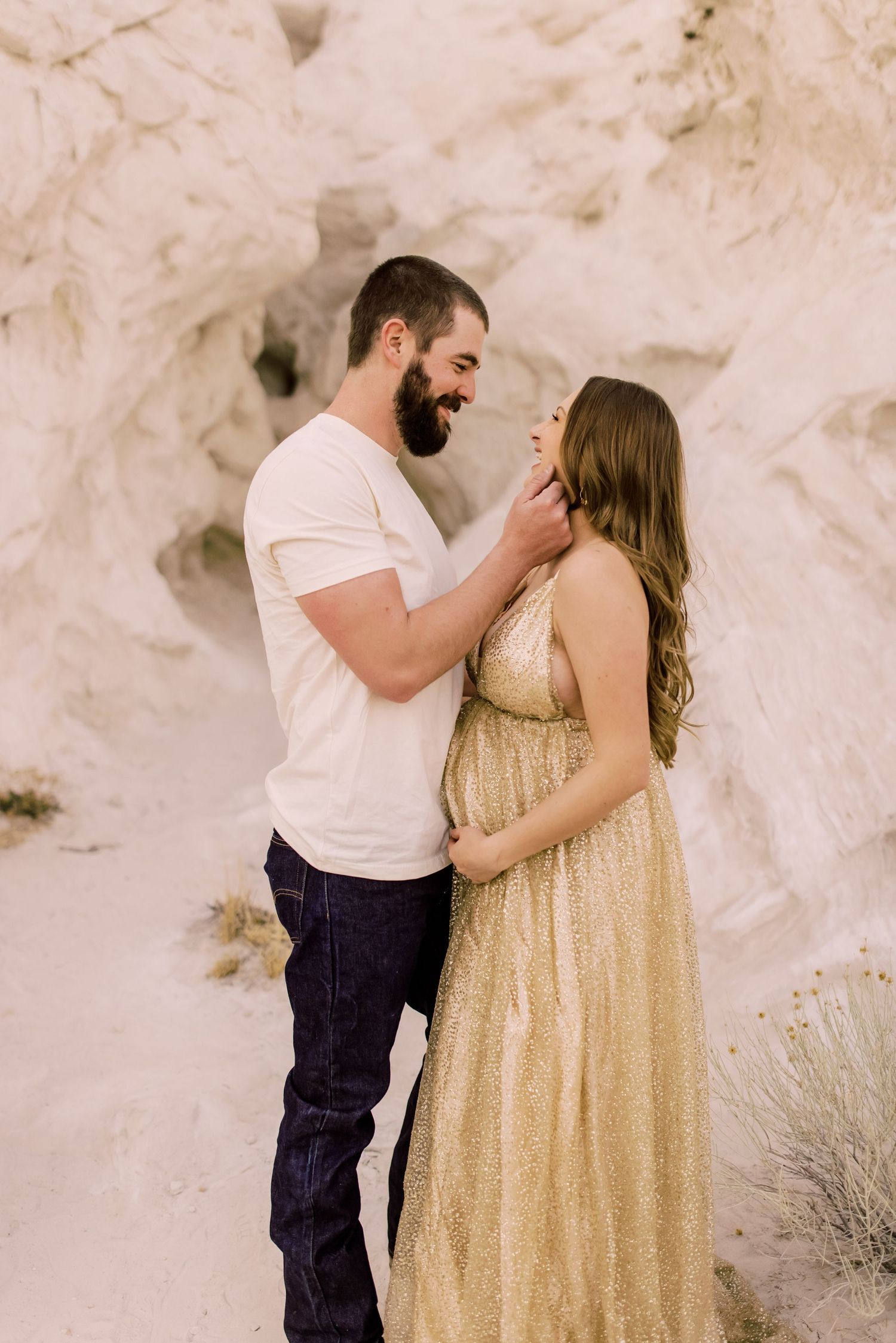 A couple shares an intimate moment against white rock formations with the woman wearing a flowing gold dress.