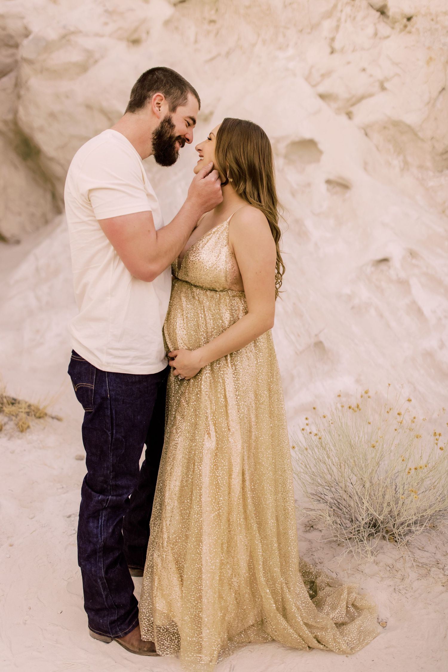 A couple shares an intimate moment against white rock formations, with the woman wearing a flowing gold sequined gown.