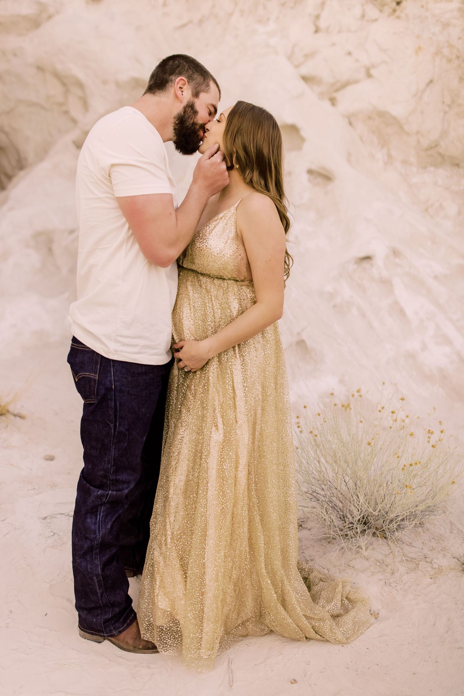 Couple sharing a tender kiss during a golden maternity photoshoot in a sandy desert landscape.