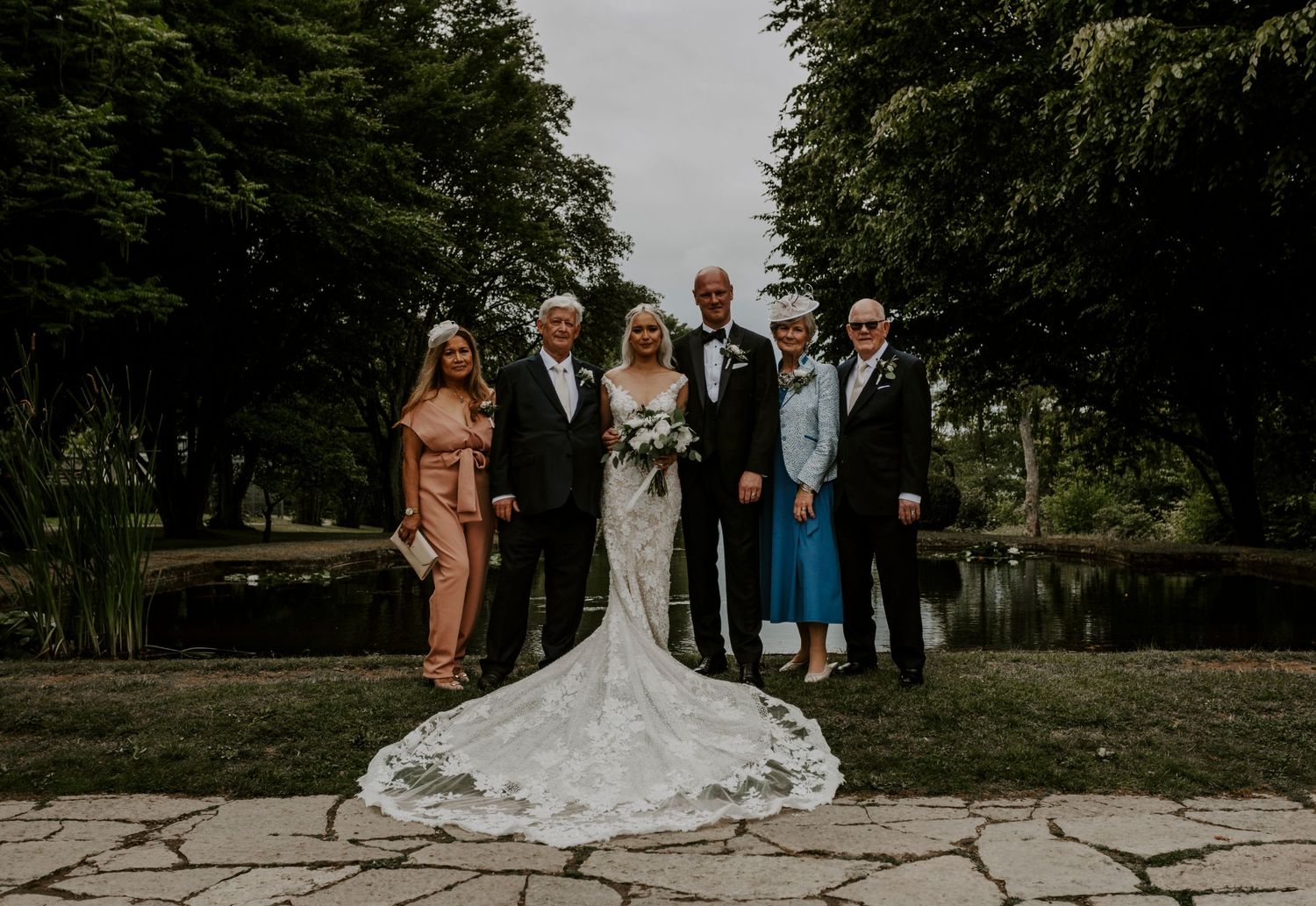 Wedding party poses for formal group photo outdoors with bride in lace gown.
