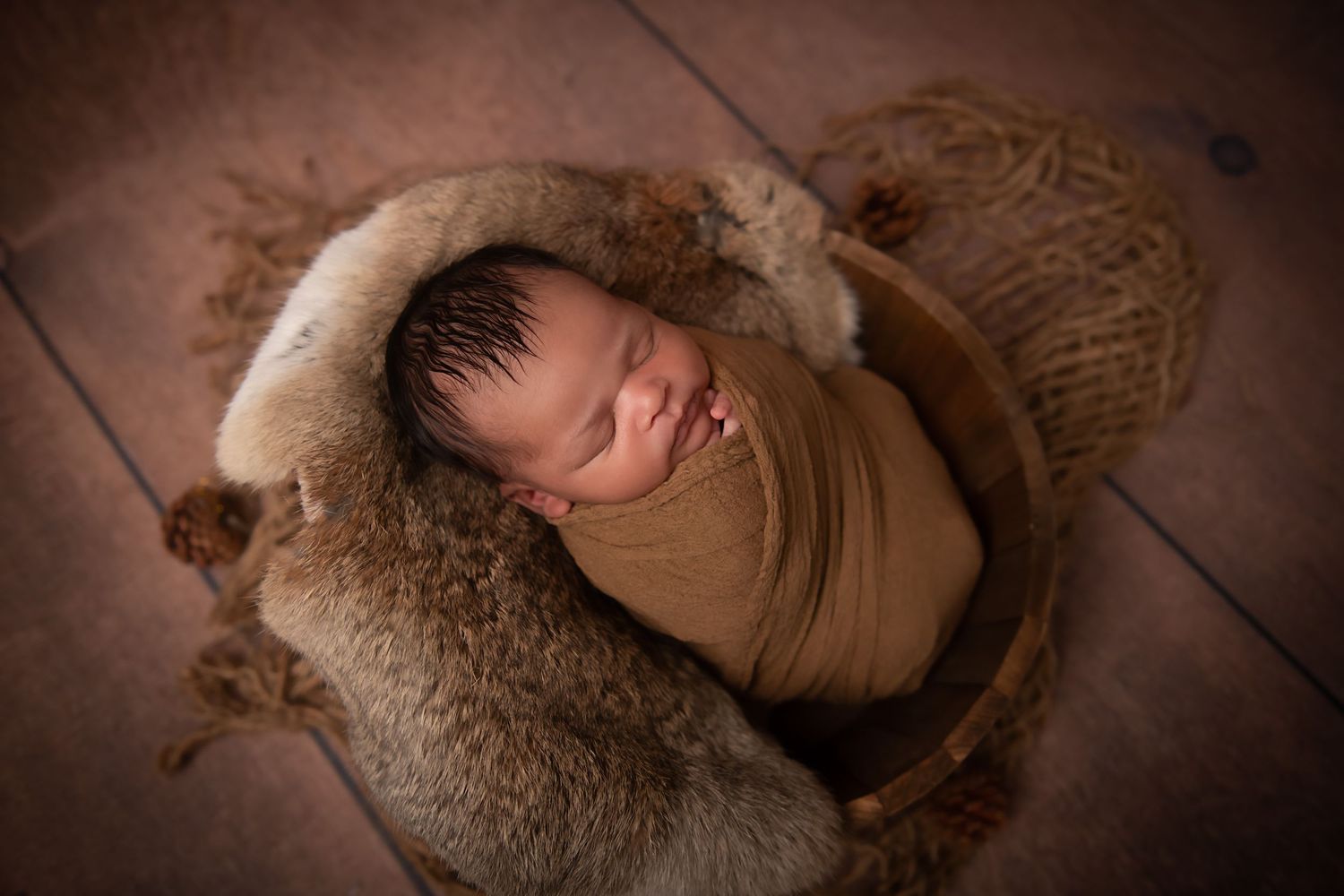 A sleeping newborn wrapped in brown fabric nestled in a cozy fur-lined basket.