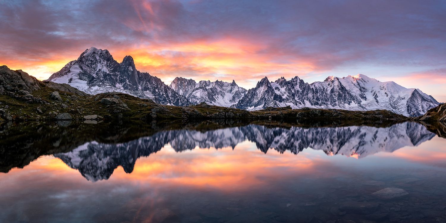 Snow-capped mountain range reflected in calm lake waters during dramatic sunset.