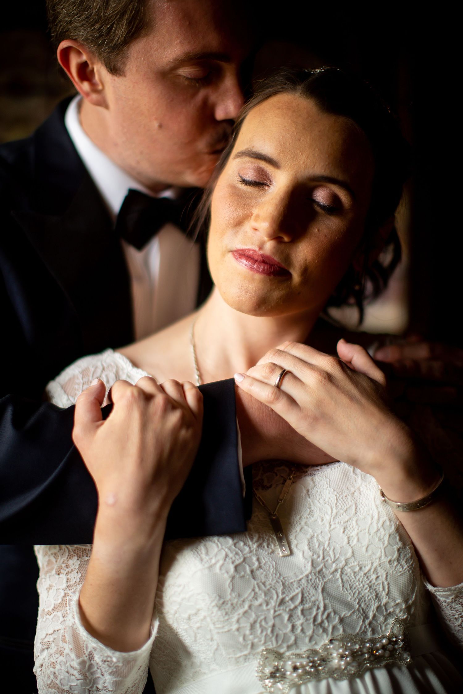 Romantic wedding portrait shows tender embrace with bride in white lace dress and groom in black tuxedo.