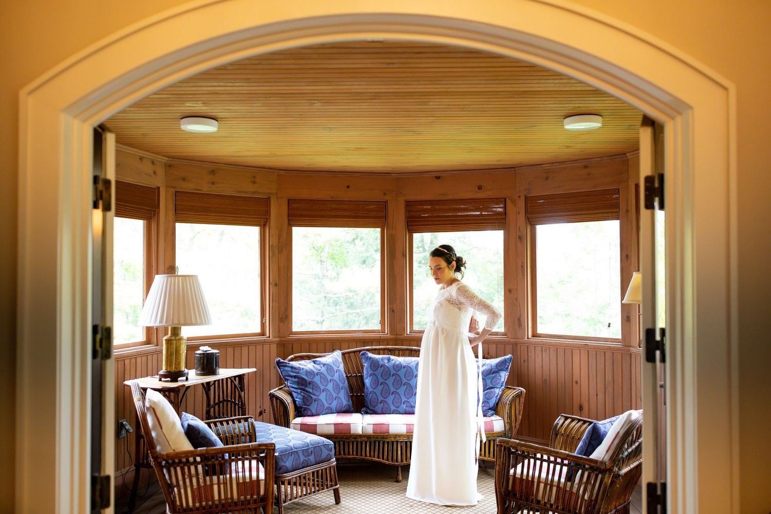A sunlit window-lined room with wicker furniture and blue cushions frames a bridal preparation moment.