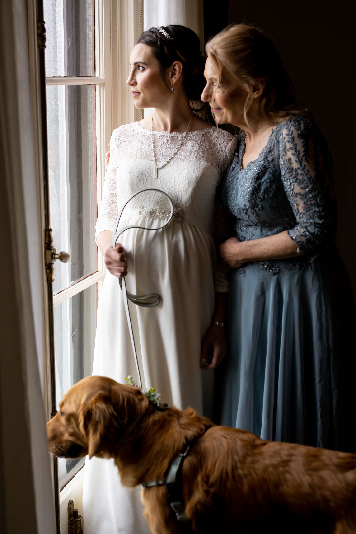 Two elegant women in formal dresses gaze out a window while a golden retriever rests nearby.