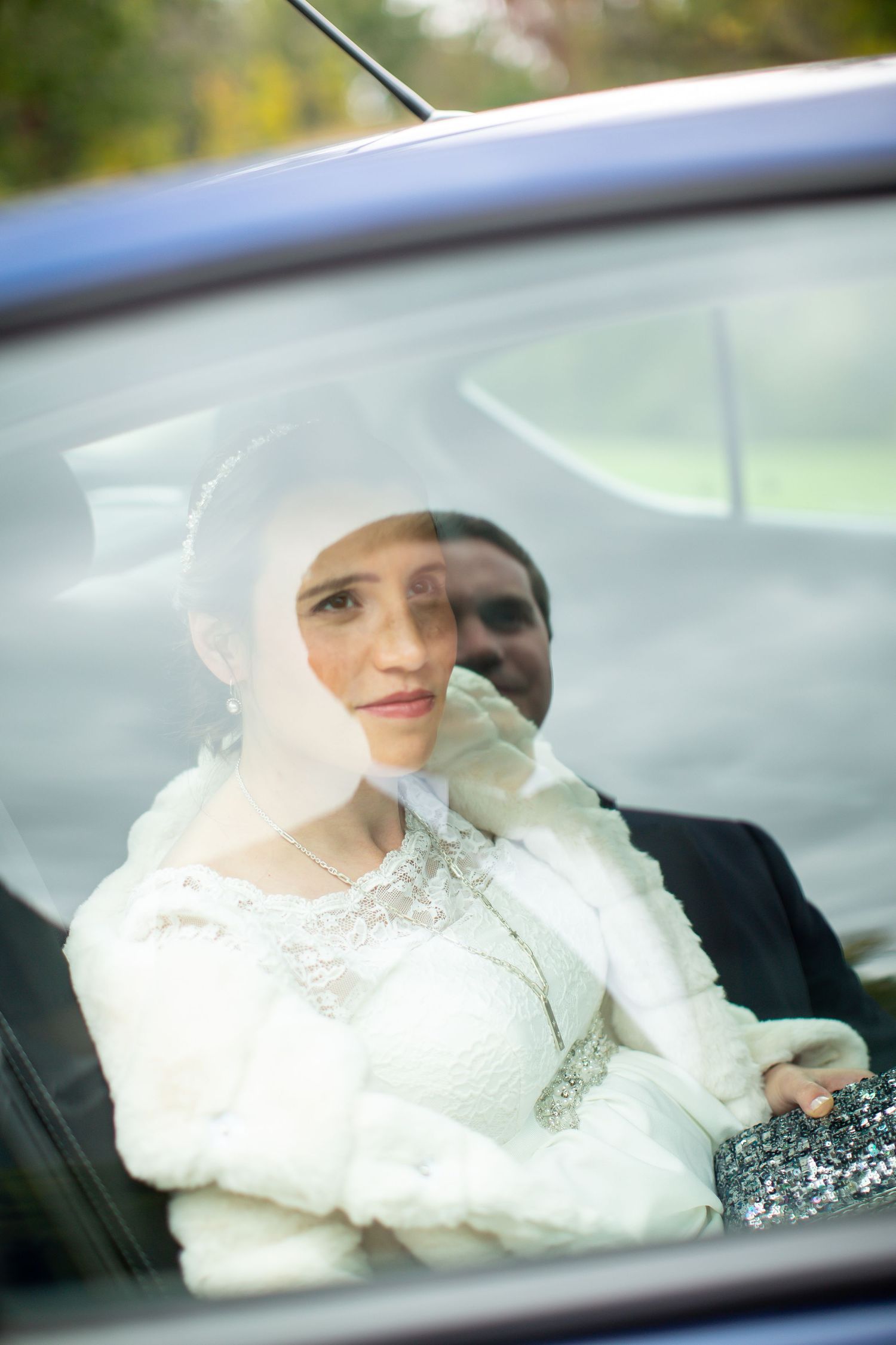 Elegant bride in white dress and fur wrap gazes through car window on wedding day.