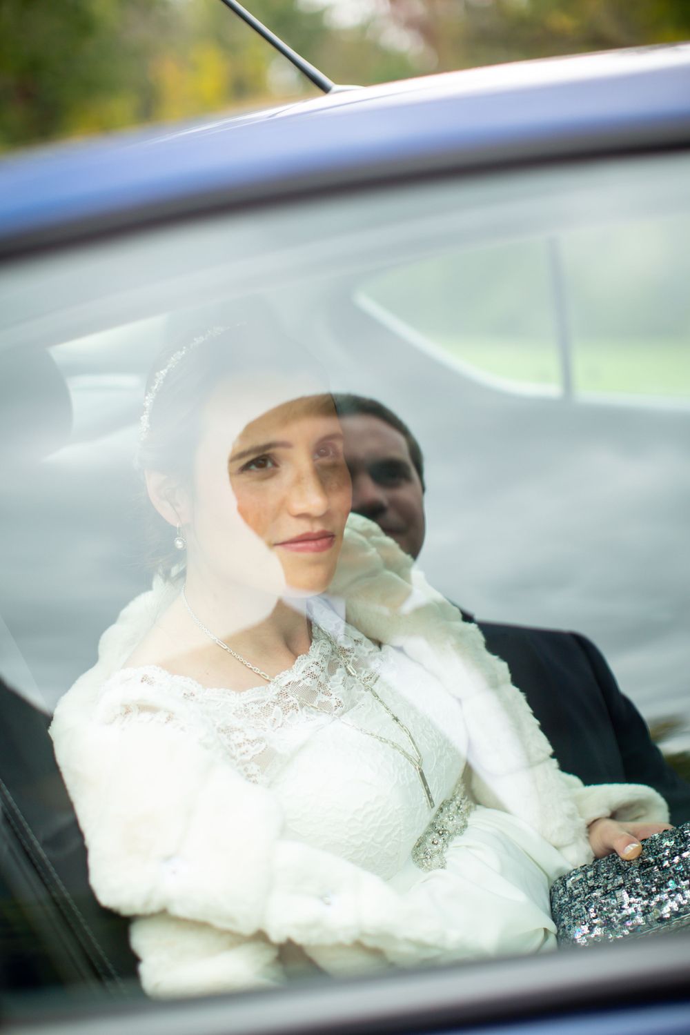 Elegant bride in white dress and fur wrap gazes through car window on wedding day.
