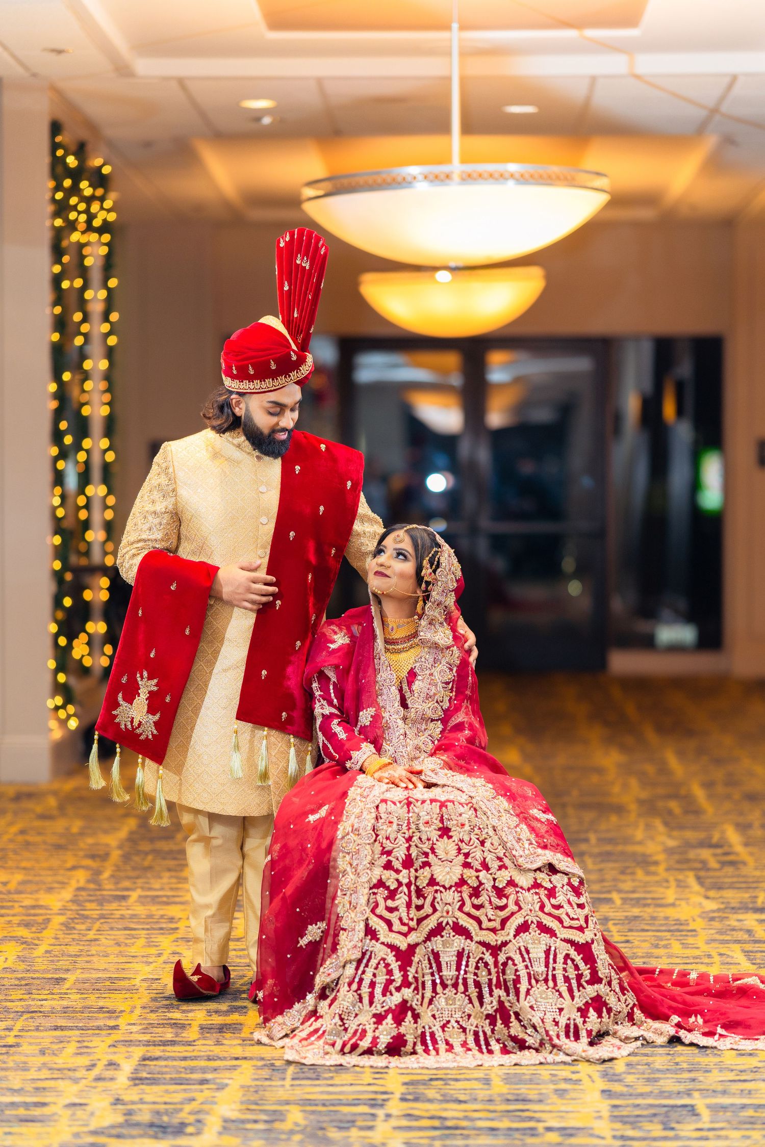 Traditional Indian bride and groom pose in elegant wedding attire with intricate red and gold details in hotel hallway.