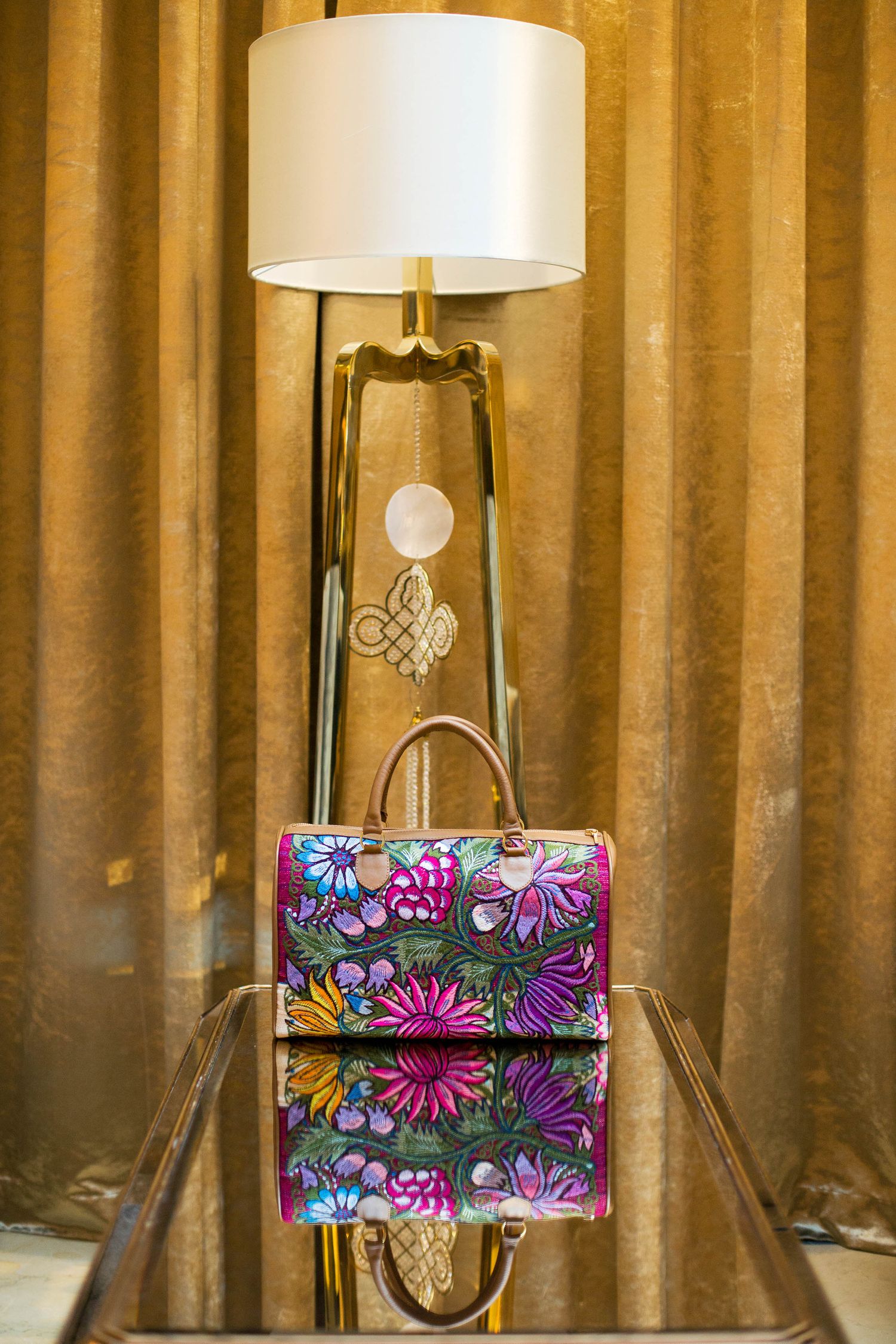 Colorful floral handbag displayed on reflective surface next to modern table lamp with gold accents.