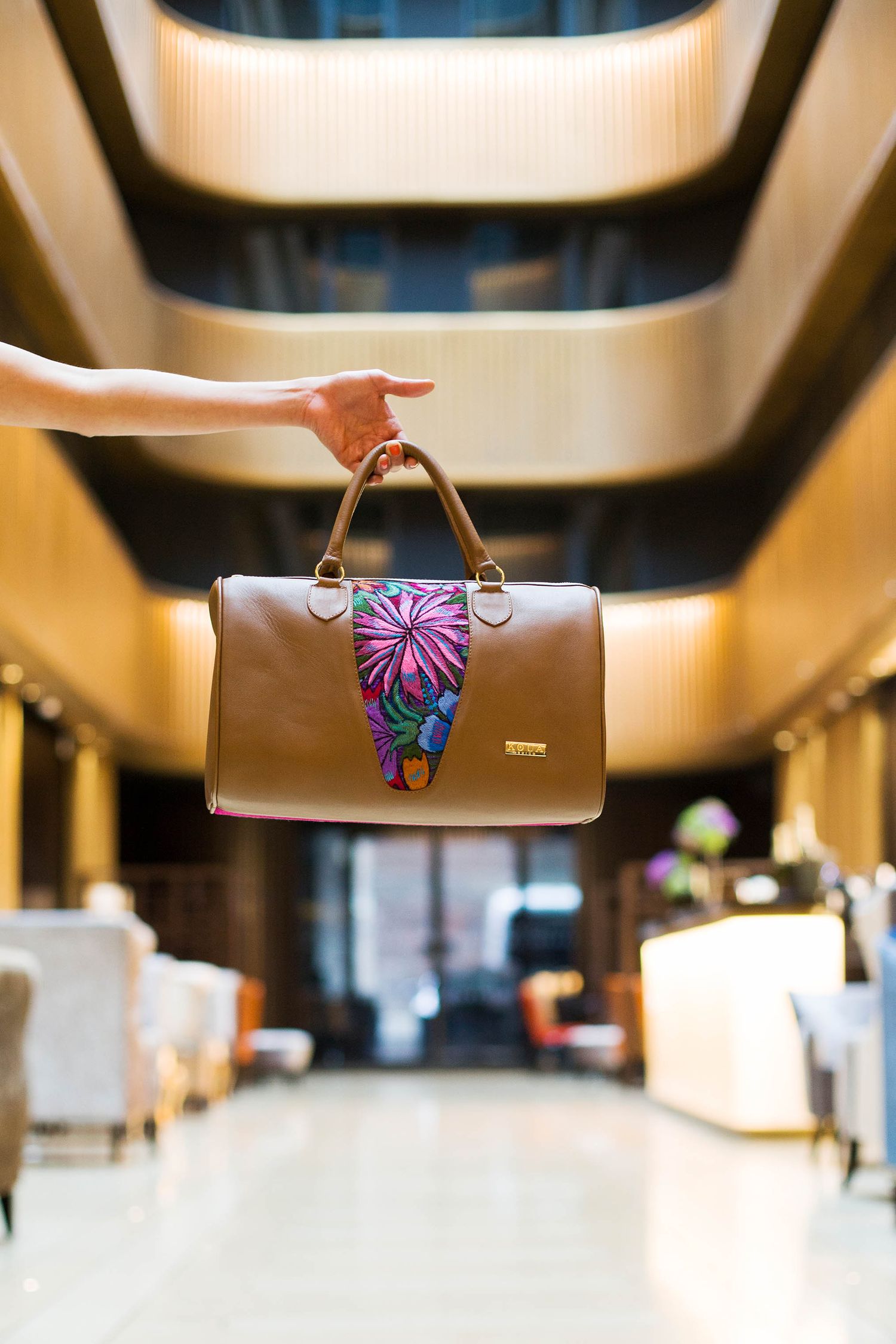 Hand holding brown leather handbag with floral pattern in elegant hotel lobby hallway.