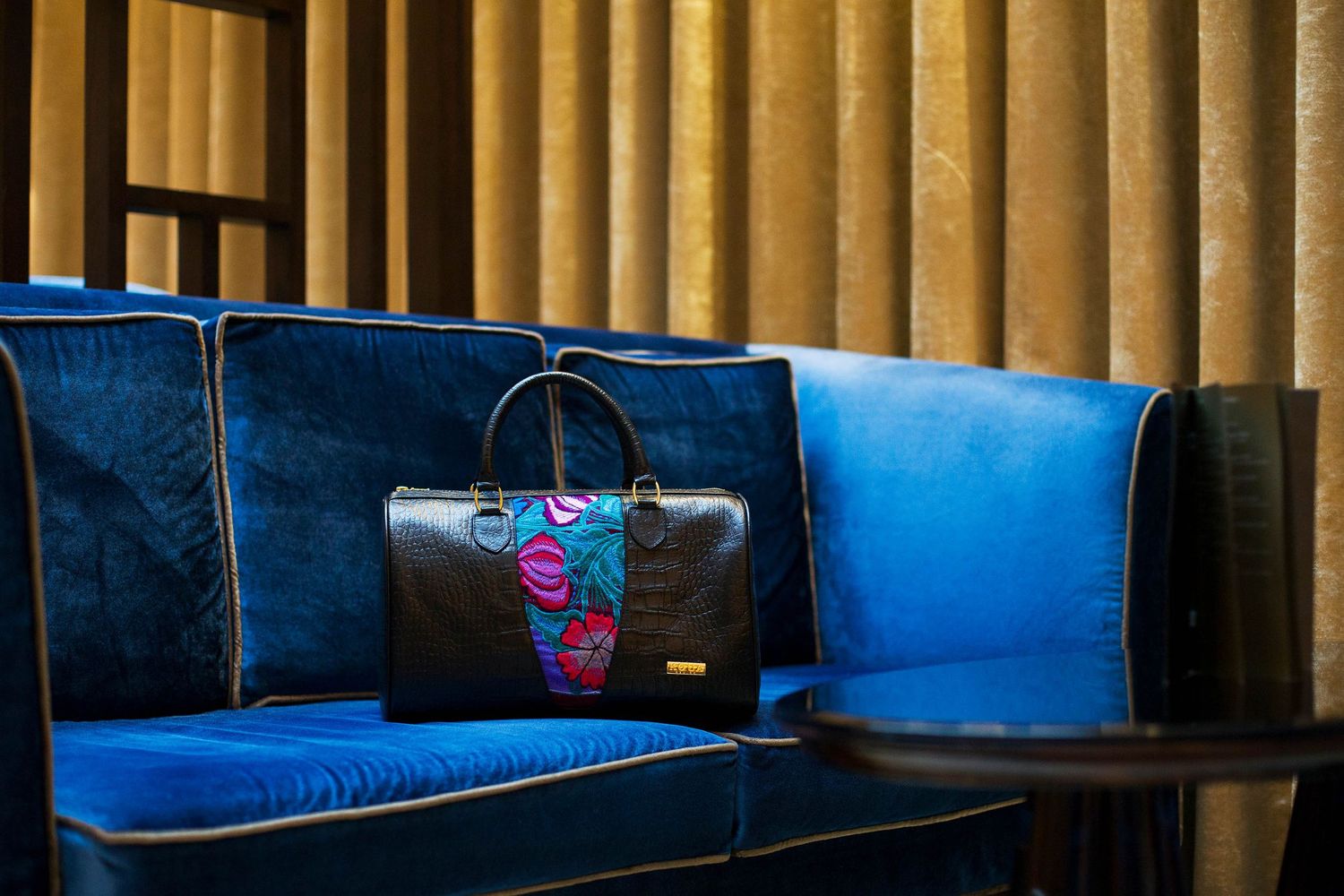 Designer handbag with colorful floral design placed on blue velvet bench seating.