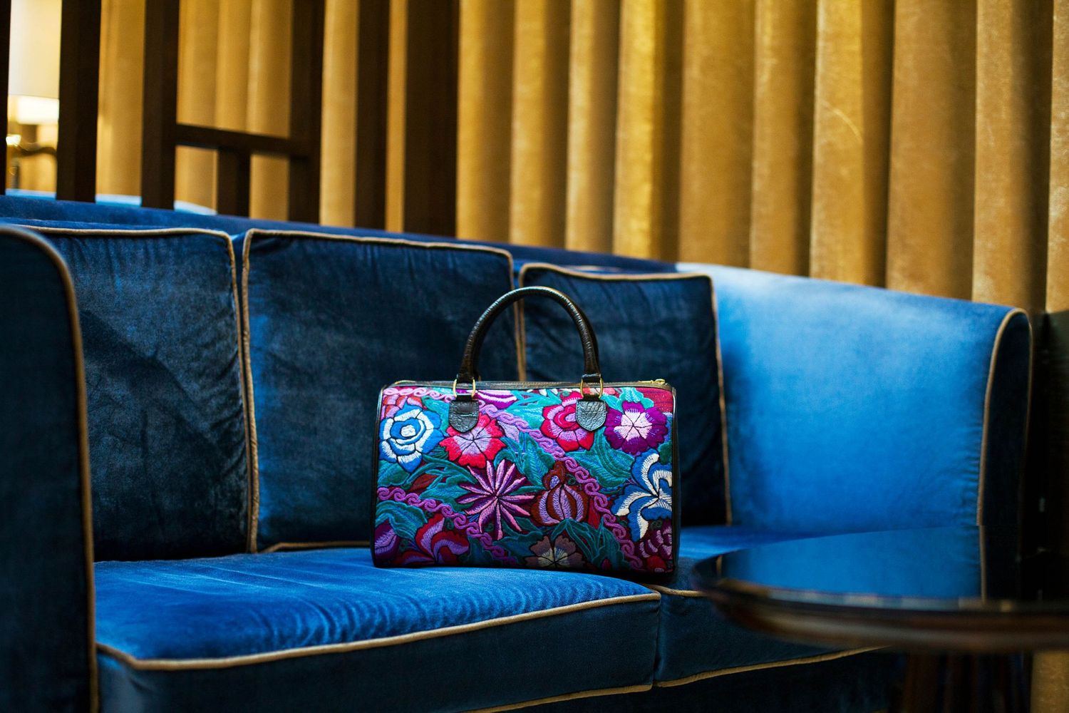 Floral print handbag displayed on blue velvet bench against gold pillared wall.