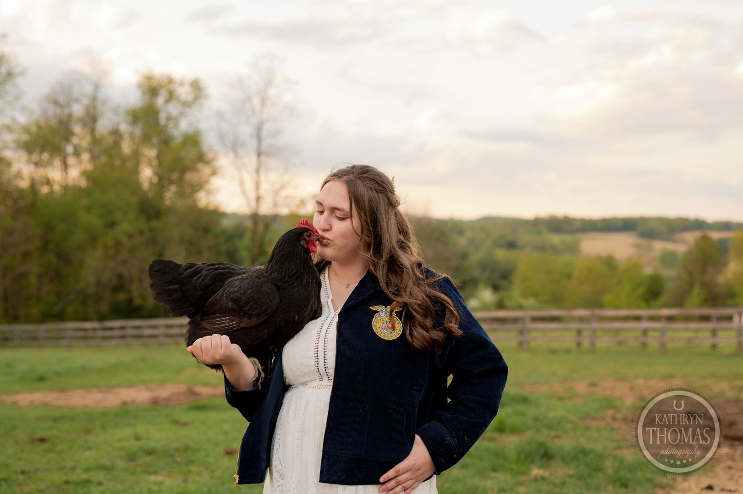 A candid outdoor portrait with a black chicken being held against a rural landscape background.