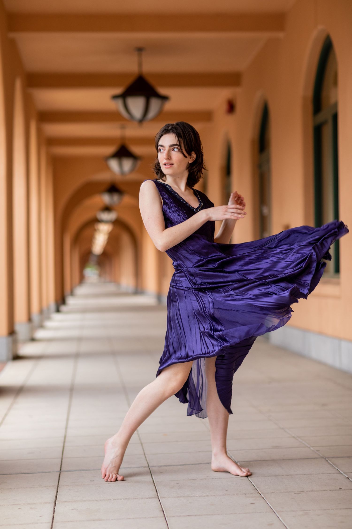 A high school senior dancer in flowing purple dress poses gracefully in arched hallway with lanterns.