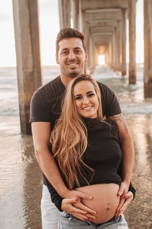 Romantic maternity photo of couple on beach under pier during golden hour.