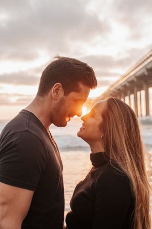 Couple sharing intimate moment at beach during golden hour with pier in background.