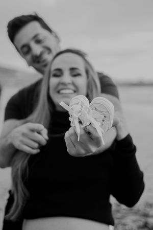 Happy couple on beach showing tiny baby shoes in black and white photography.