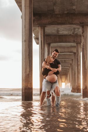Couple sharing intimate moment during beach maternity session under wooden pier structure.