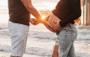 Silhouetted couple holding hands at sunset on beach with pregnant belly visible.