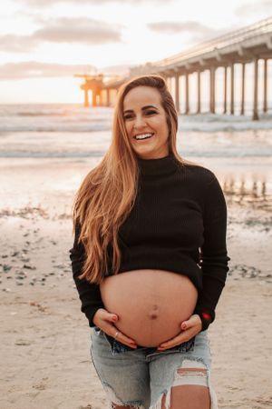 Expecting mother in black sweater smiling at beach during sunset.