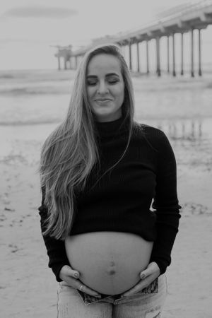 Black and white maternity portrait at beach with pier in background.