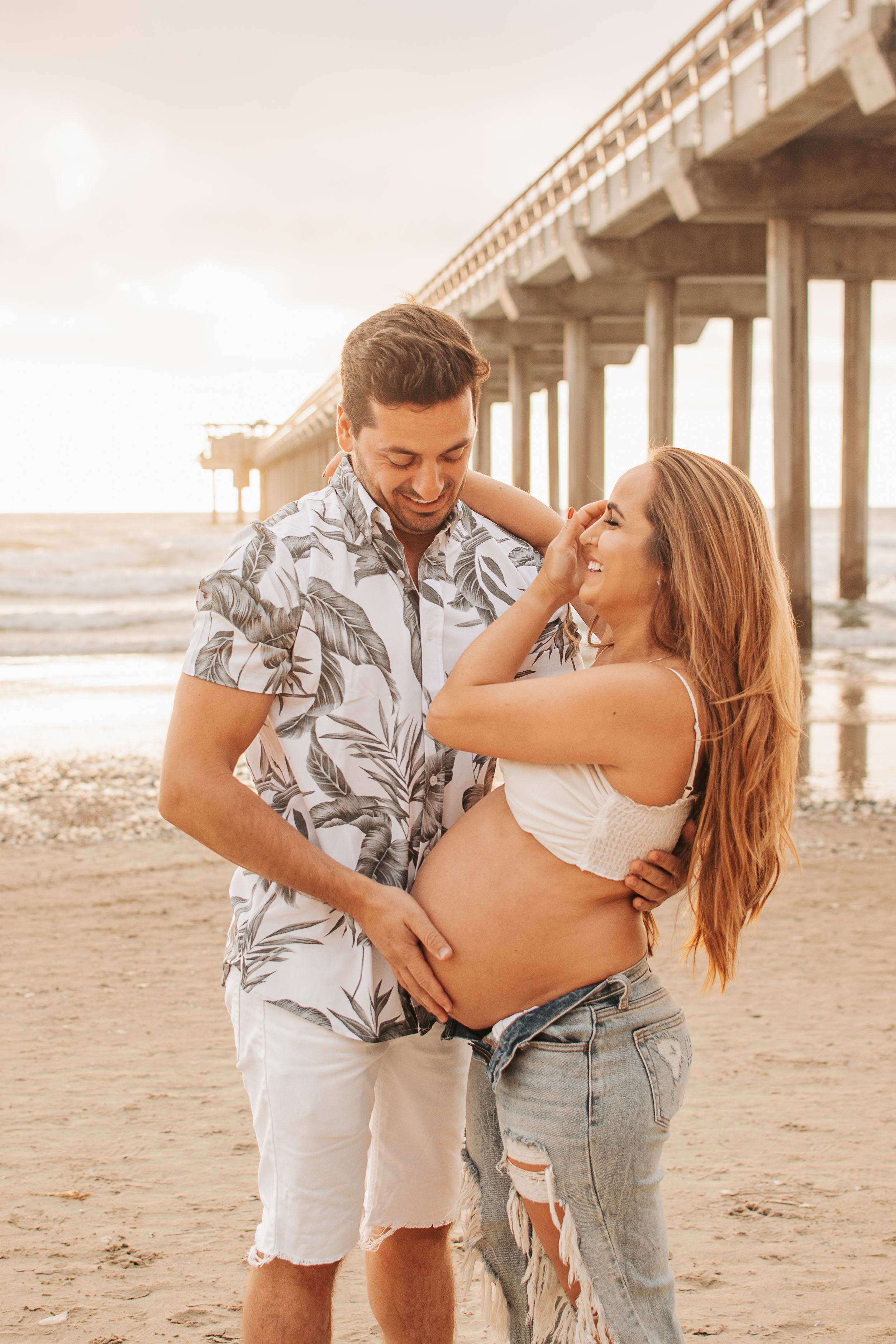Romantic sunset beach photo of couple by pier during maternity photoshoot.