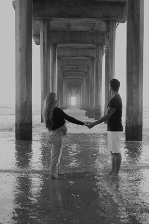 Black and white photo of couple holding hands under pier at beach with dramatic vanishing point perspective.