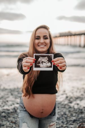 A pregnancy announcement showing an ultrasound photo held up at a beach with a pier in the background.