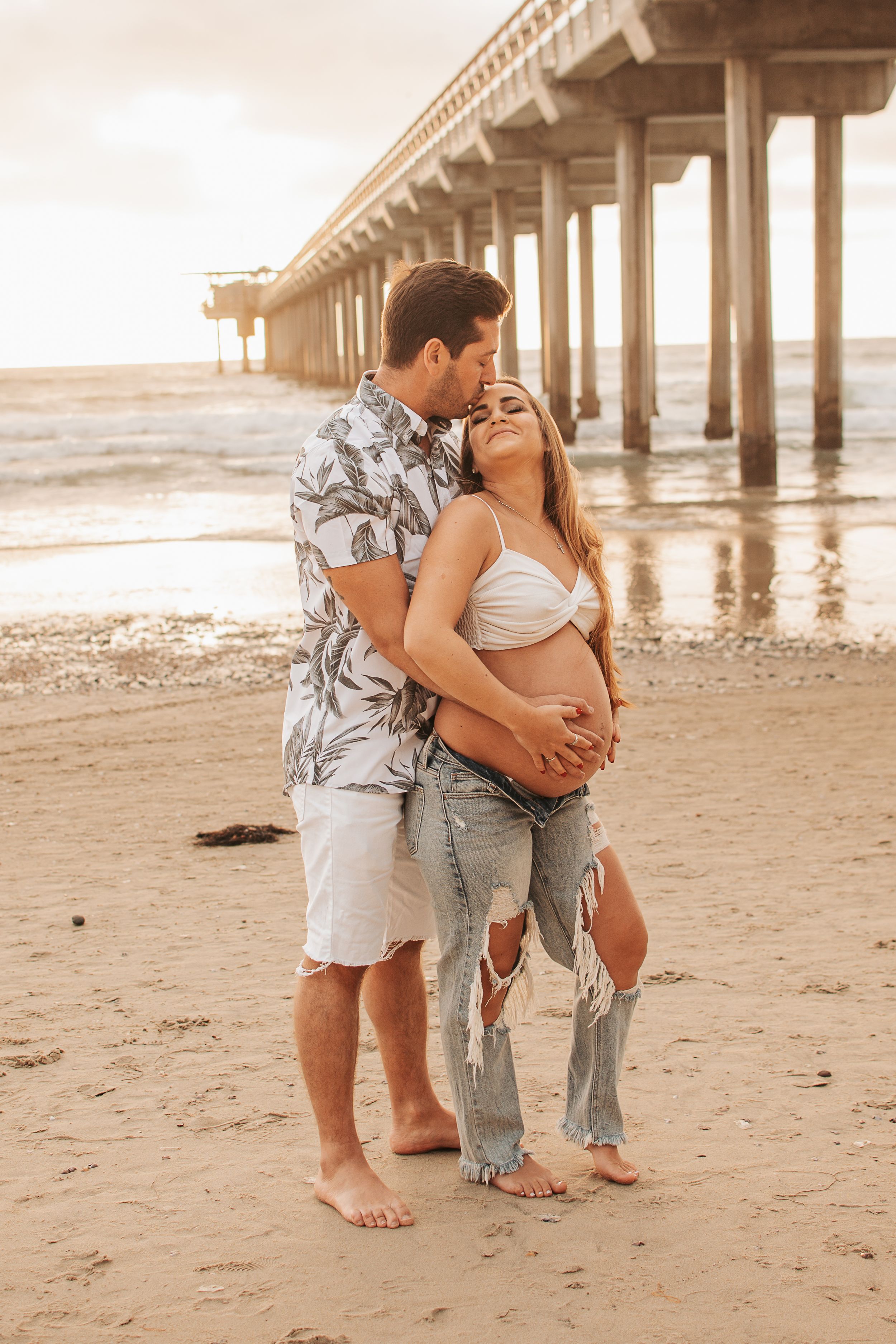 Loving couple embracing on beach near pier during golden hour maternity session.