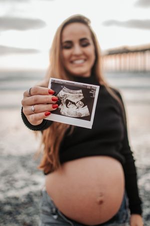 Expectant mother holding ultrasound photo at beach during sunset.