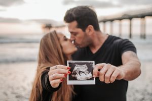 Couple sharing a kiss while holding up an ultrasound photo on the beach.