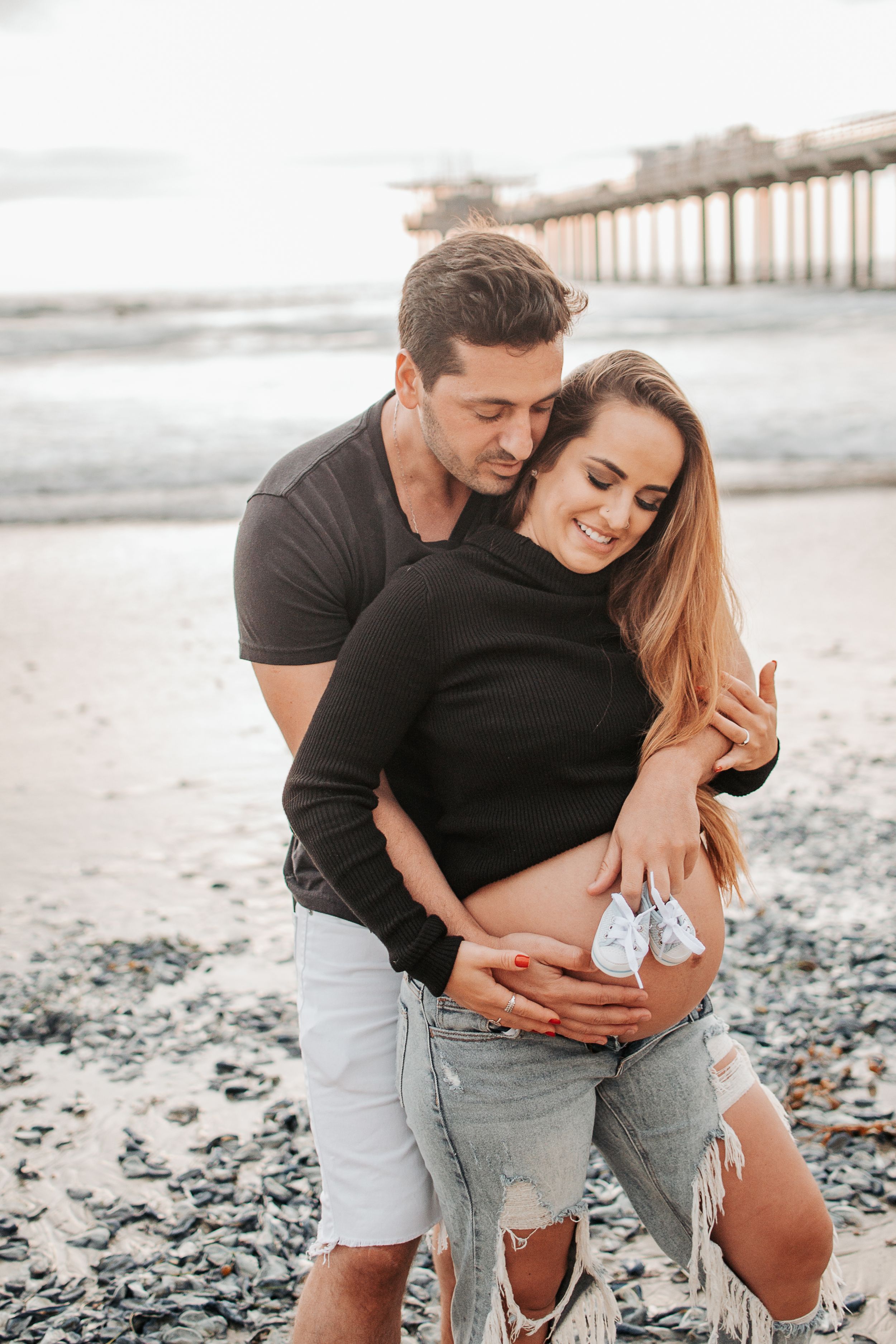 Romantic beach maternity photo with couple holding baby shoes.