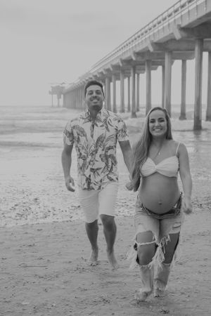 Black and white photo of couple walking together under pier on sandy beach.