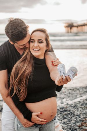Expecting couple embracing on rocky beach while holding baby shoes.
