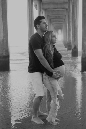 Black and white photo of couple standing in water under pier.