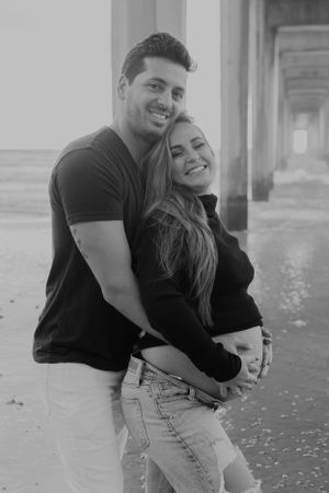 Black and white portrait of couple embracing under beach pier.