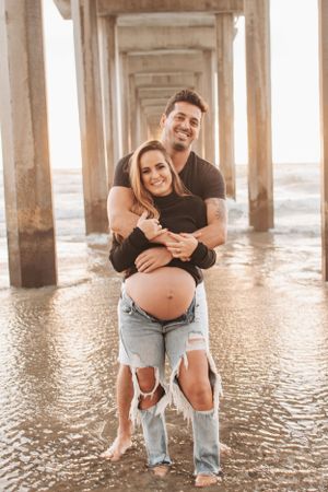 Expectant couple embracing under pier supports during maternity photoshoot at beach.