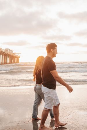 Couple walking barefoot on beach near pier during sunset.